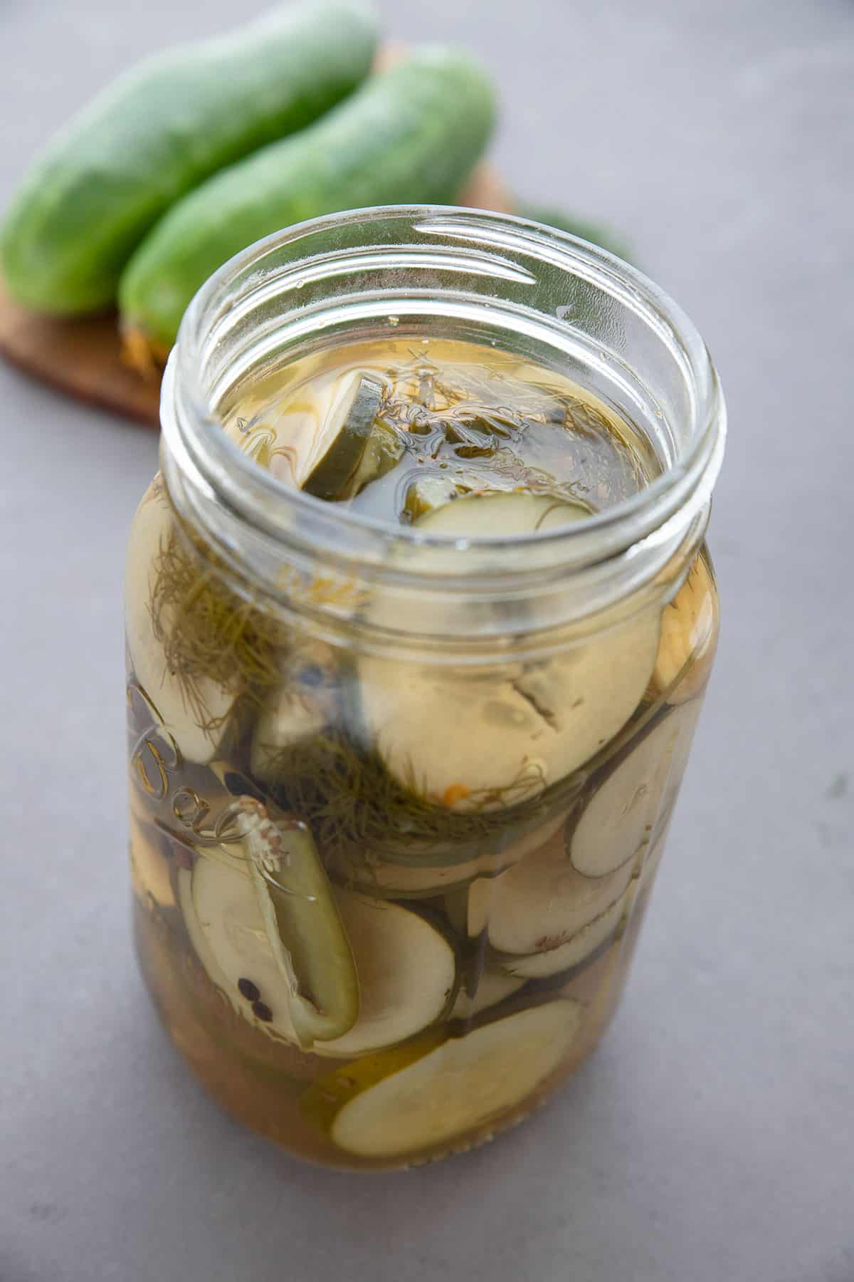 A jar of keto refrigerator pickles in front of a cutting board with cucumbers on it.