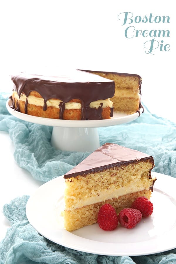Keto Boston Cream Pie recipe - a slice of the cake on white plate with raspberries, with the rest of the cake on a white cake stand in the background