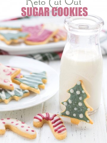 Keto sugar cookies on a white table with a bottle of milk. More sugar cookies in the background, all decorated with sugar-free royal icing