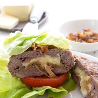 A keto stuffed burger in a lettuce wrap on a white plate, with brie and caramelized onions in the background.