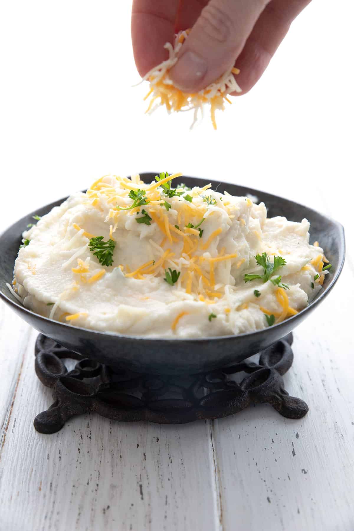 A bowl of creamy mashed cauliflower in a black bowl, with grated cheddar being sprinkled over top. 
