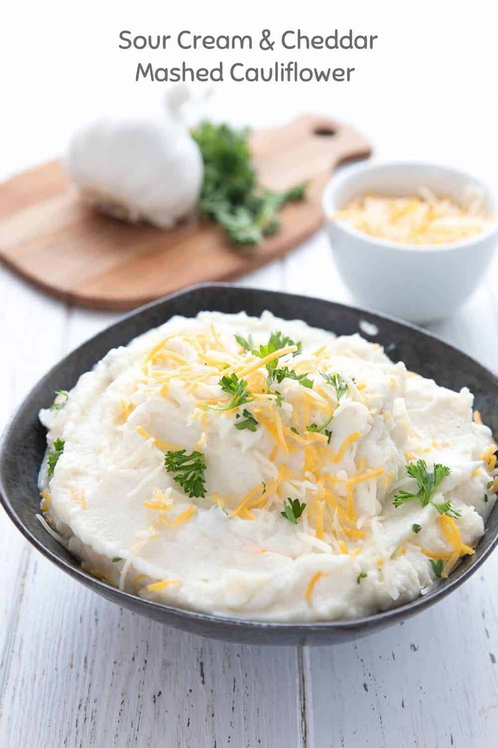 Titled image of keto mashed cauliflower in a black bowl, topped with cheddar and parsley.