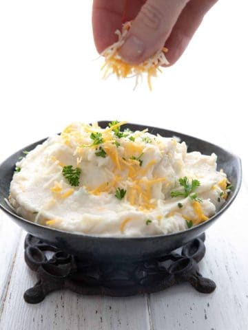 A bowl of creamy mashed cauliflower in a black bowl, with grated cheddar being sprinkled over top.