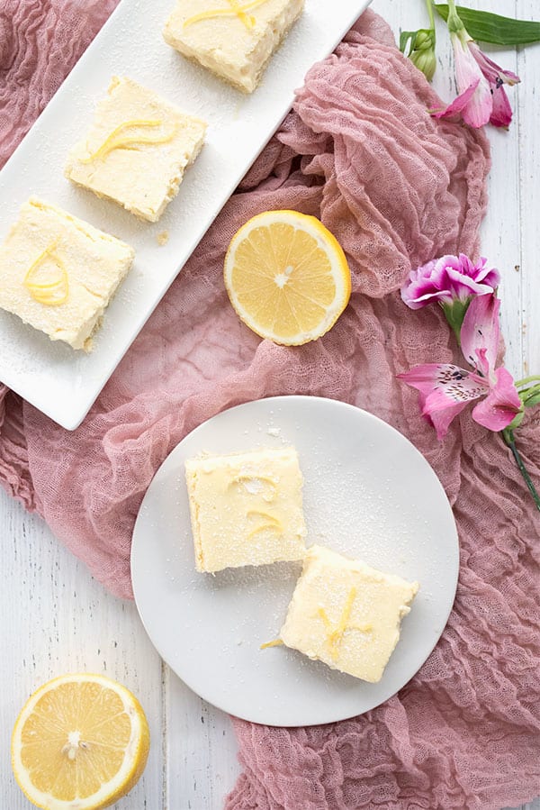 Top down photo of lemon cheesecake bars on two white plates with a rose colored cloth and cut lemons. 