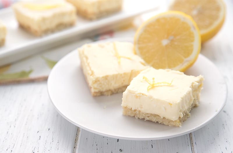 Two lemon cheesecake bars on a white plate with cut lemons in behind. 