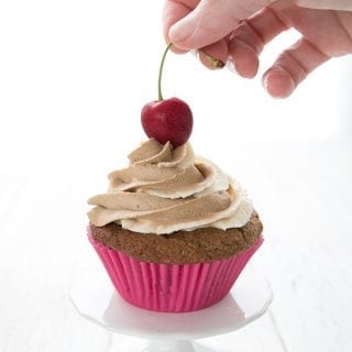 A hand placing a cherry on top of a root beer cupcake.