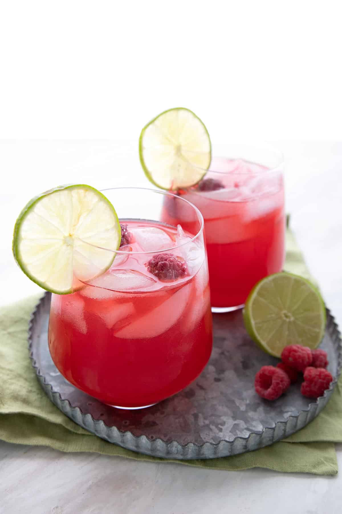 Two glasses of raspberry limeade on a pewter platter with fresh lime and berries.