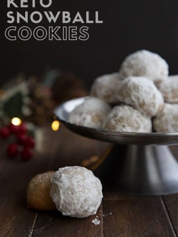 Titled image of keto snowball cookies on a metal cake stand with holiday lights in the background.