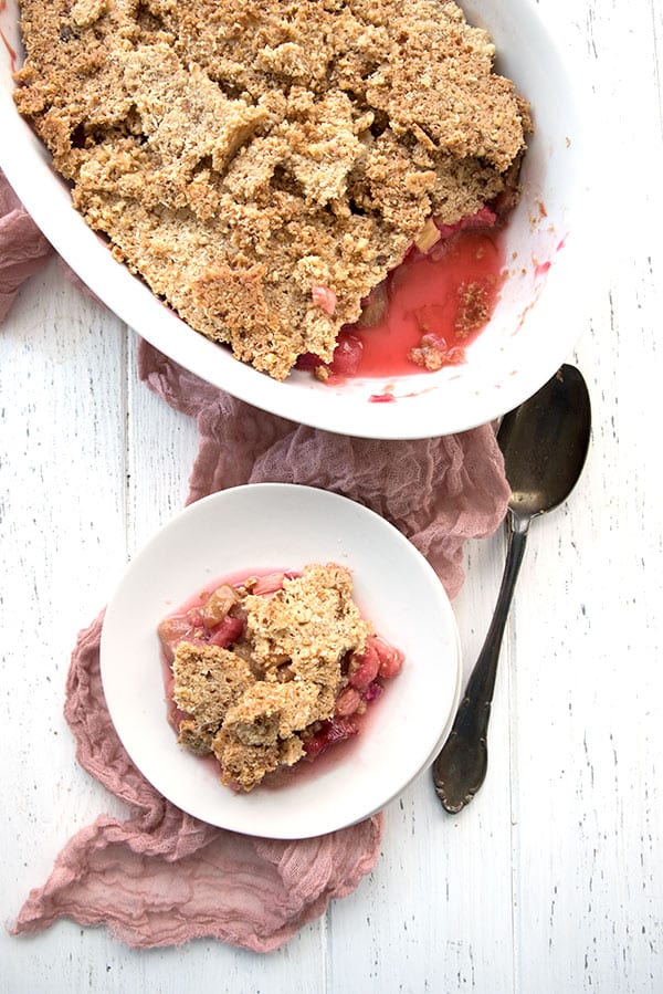 Top down photo of rhubarb crisp on a white table. A white plate with one serving beside the main oval dish.