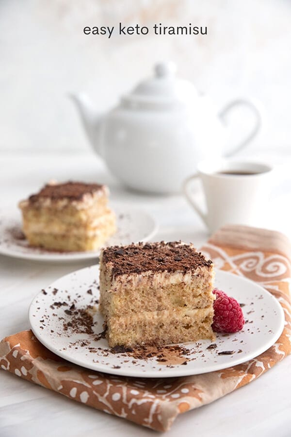 Titled image of keto tiramisu on white plates over a patterned napkin, with a white coffee cup and coffee pot in the background.
