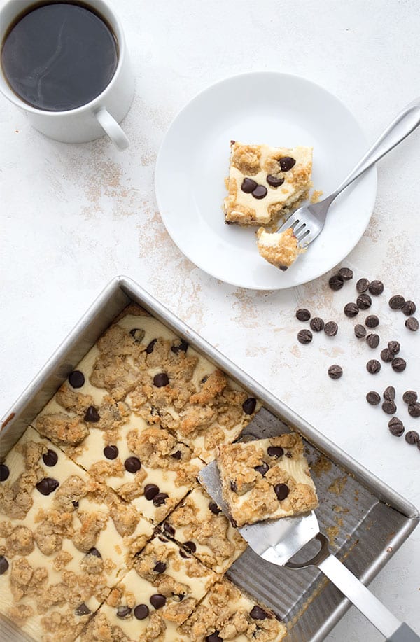 Top down photo of chocolate chip cookie cheesecake bars in a pan, and a white plate with one bar on it, along with a cup of coffee. 