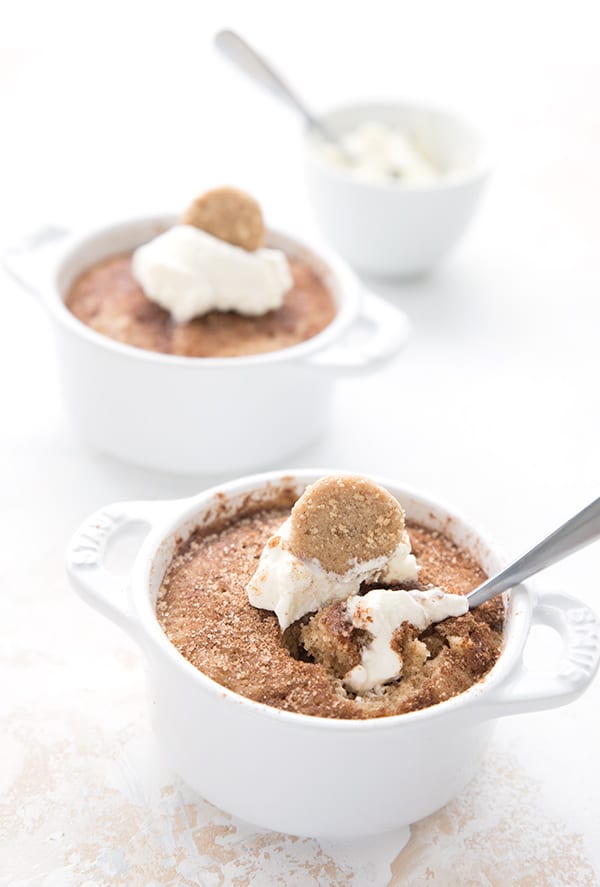 Two snickerdoodle mug cakes on a white table with a bowl of whipped cream in the background. 
