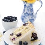 A slice of lemon blueberry zucchini later cake on a white plate with a vase of yellow flowers in the background.