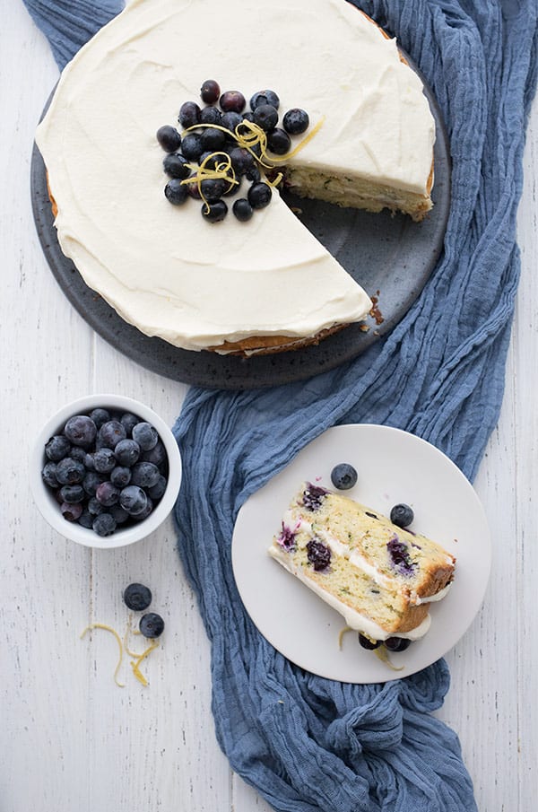 Top down photo of keto lemon blueberry cake. A slice of cake on a white plate, with the rest of the cake on a blue plate, over a blue cloth. A bowl of blueberries to the side. 