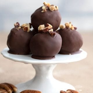 Titled image of keto pecan pie truffles on a small white cake stand with pecans strewn around.
