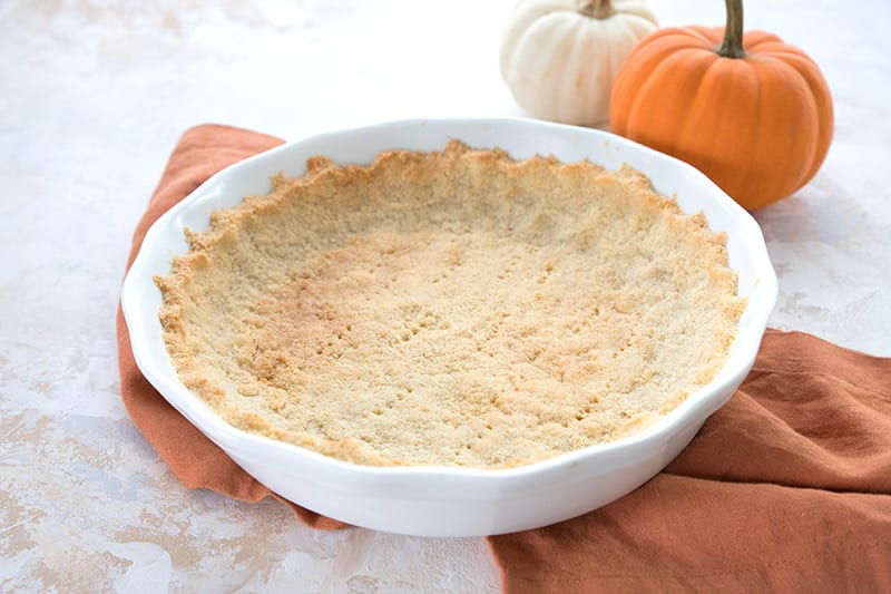 A baked low carb pie crust on an orange napkin with small pumpkins in the background. 