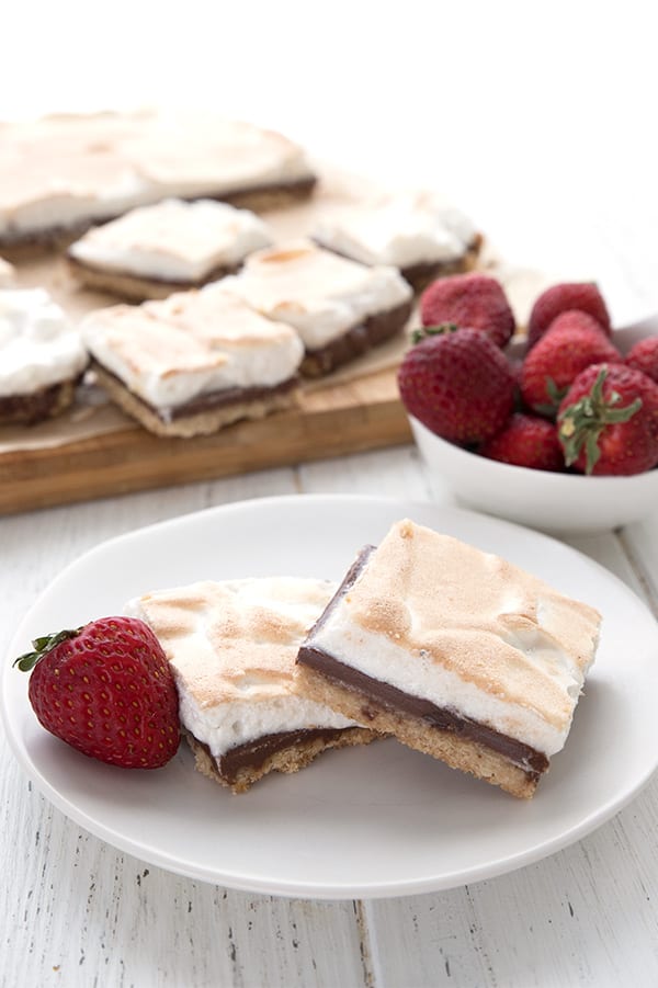 Two keto smores bars on a white plate with a strawberry. A bowl of strawberries and a cutting board filled with more bars in the background.