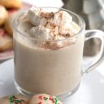 A mug of keto eggnog with whipped cream on top in front of a plate of cookies.