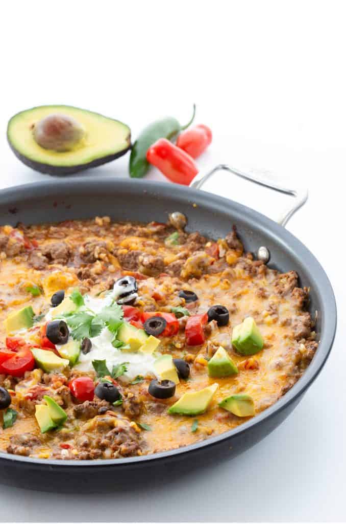 A pan filled with keto Mexican style cauliflower rice on a white background, with avocado, jalapeno, and tomatoes in the background. 