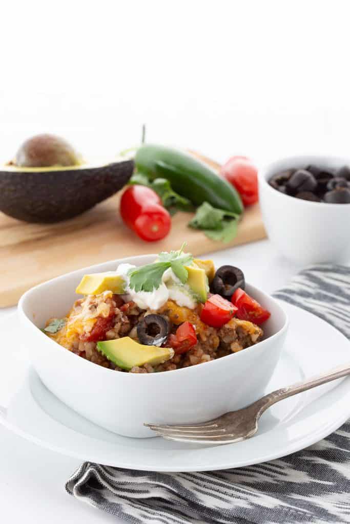 A bowl of Mexican Cauliflower Rice with avocado, tomatoes, jalapeños, and olives on a cutting board in the background. 