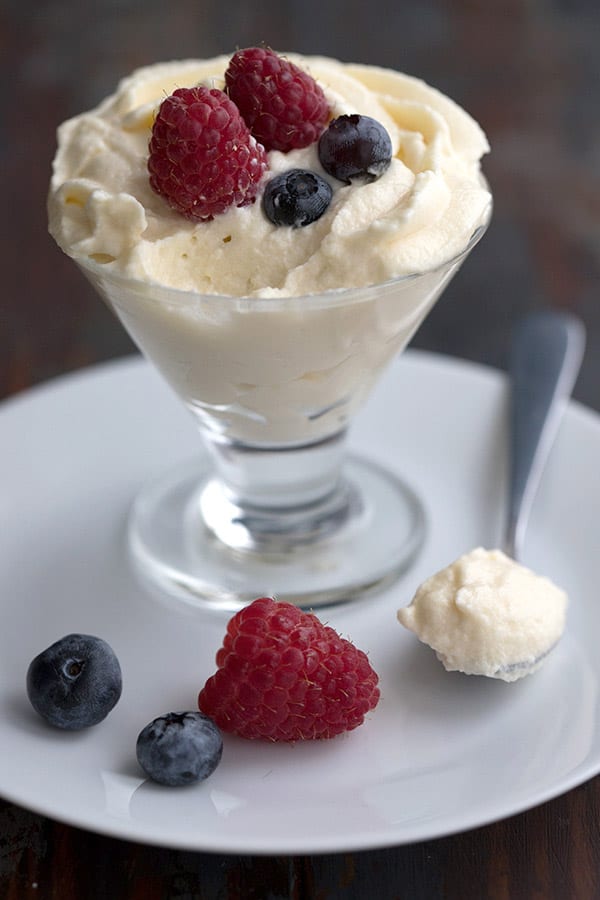 A cup of keto white chocolate cheesecake mousse sits on a white plate, with berries on top and scattered around. A spoonful of the mousse sites beside the cup. 
