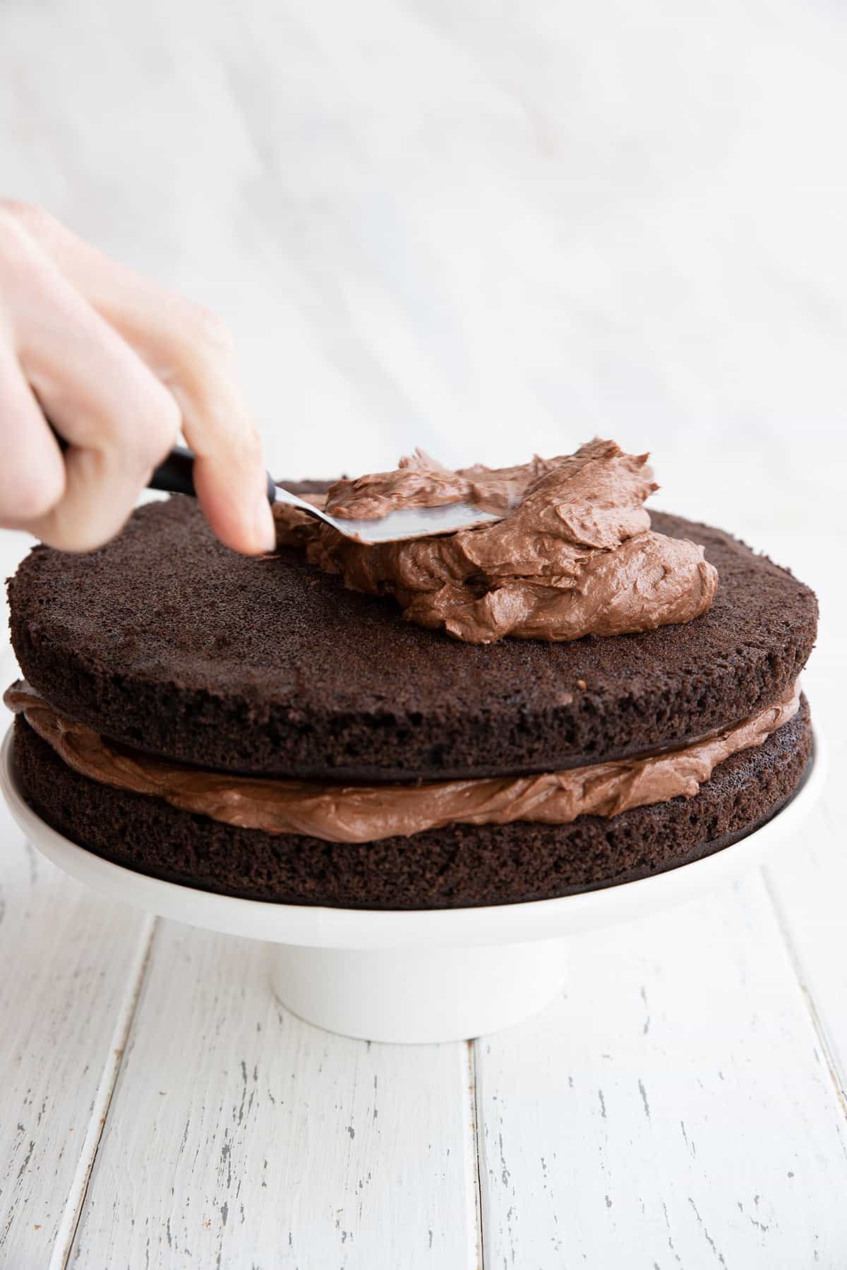 A chocolate cake on a white cake stand being spread with keto chocolate frosting. 