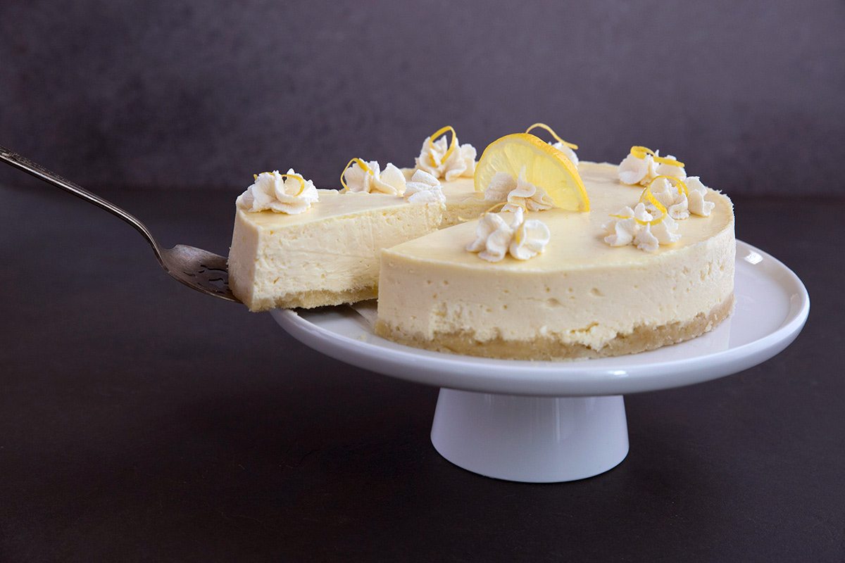 A slice of low carb lemon cheesecake being removed from the whole on a cake stand