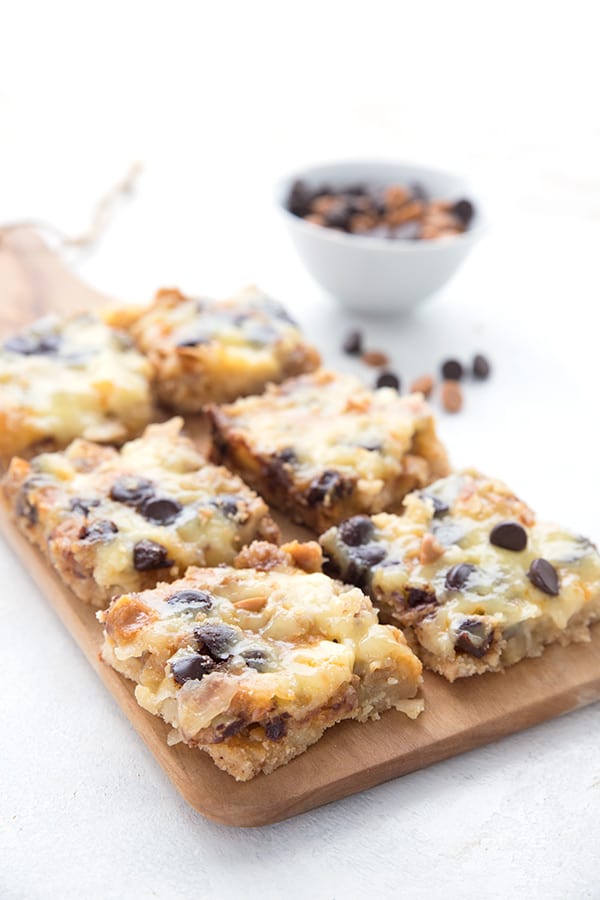Keto magic cookie bars laid out in rows on a wooden cutting board, with a bowl of chocolate and butterscotch chips in the background. 