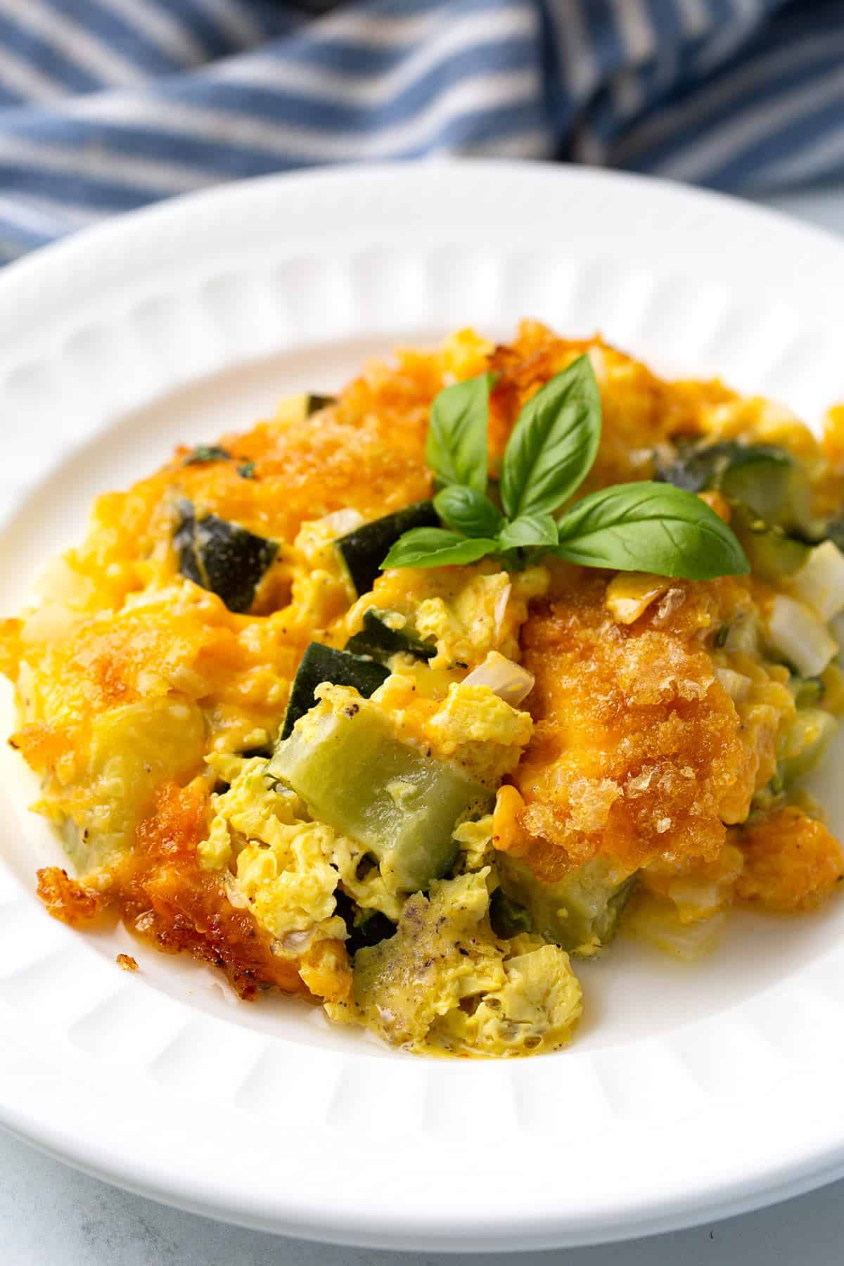 A serving of Zucchini Casserole on a white dish with a sprig of basil on top. 