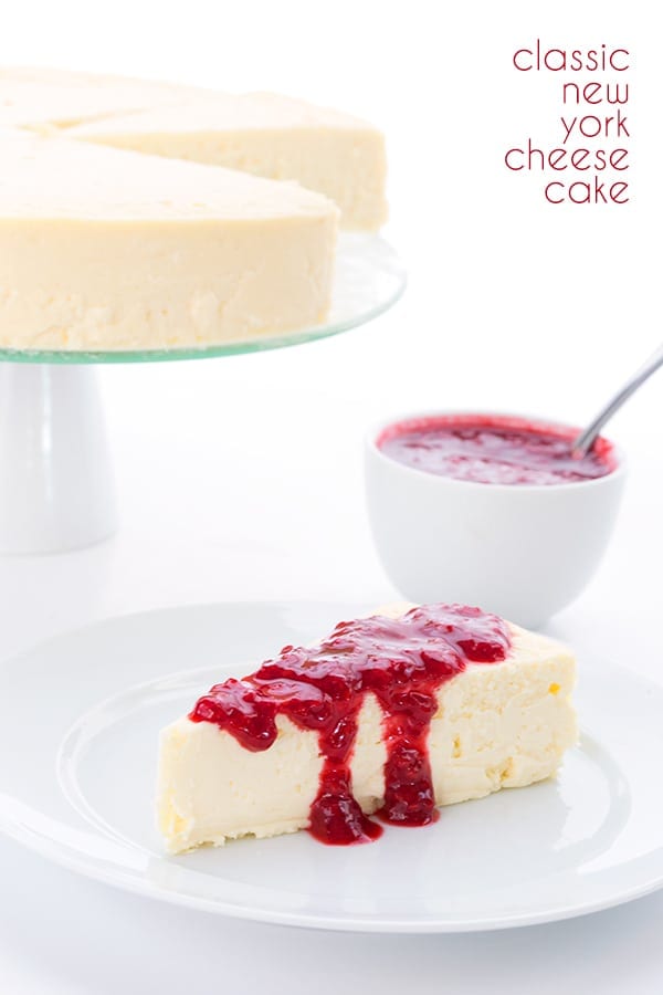 Keto cheesecake on a white plate with sugar free raspberry sauce. In the background is the full cheesecake on a cake stand and a bowl of the sauce