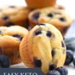 Titled Pinterest image of a keto blueberry muffin on a white table with fresh blueberries strewn around.