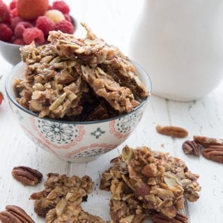 A bowl of granola clusters with berries in the background