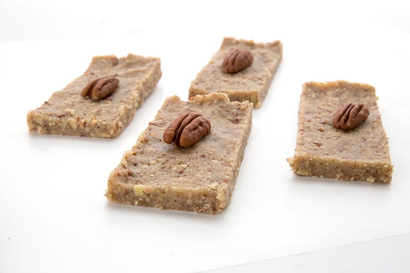 Four pecan pie bars on a white table, each with a pecan on top. 