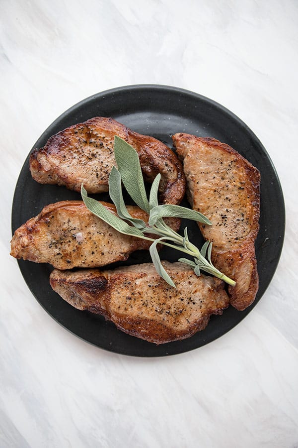 Top down photo of keto air fryer pork chops on a black plate with a sprig of sage on top. 