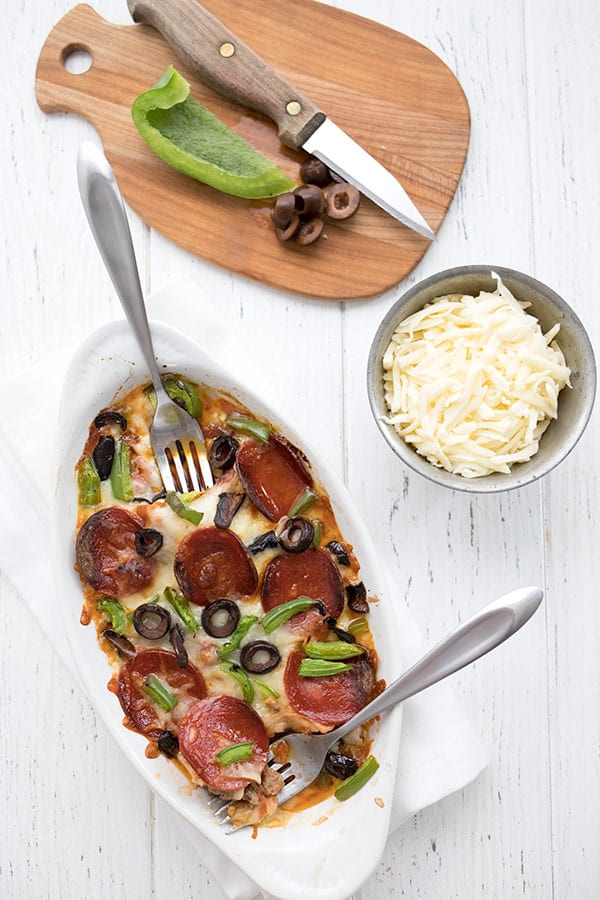 Top down foto van korstloze pizza voor twee met een kom versnipperde kaas en een snijplank met groene peper en zwarte olijven erop.