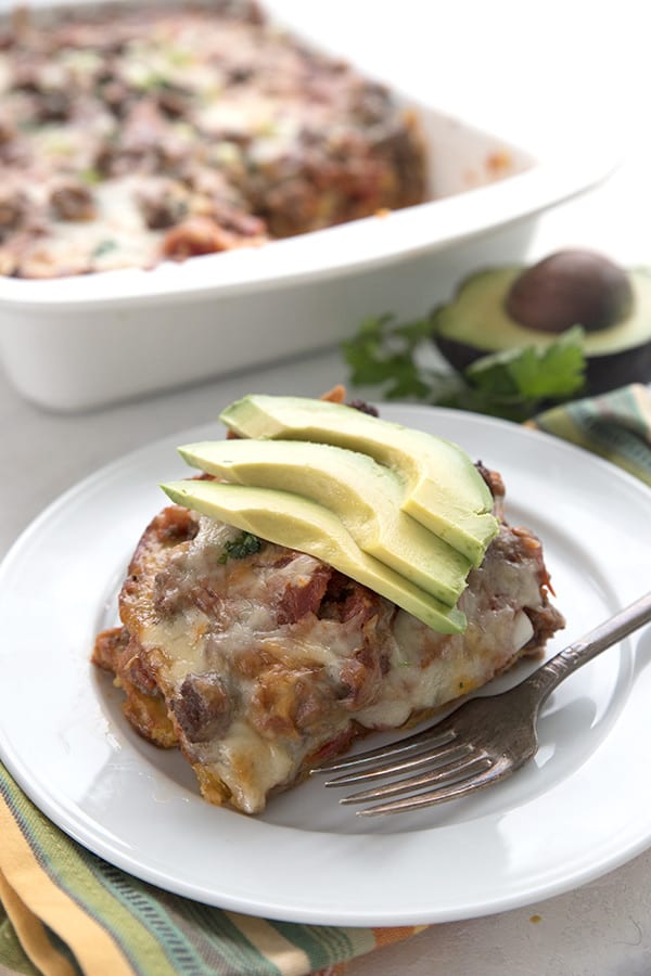 Beef enchilada casserole on a white plate in front of the pan. Slices of avocado on top. 