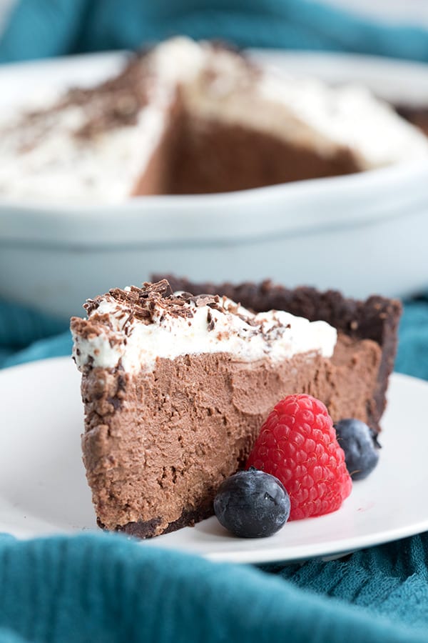 A slice of chocolate mousse pie on a white plate over a blue tablecloth. Fresh berries on the plate beside the pie. 