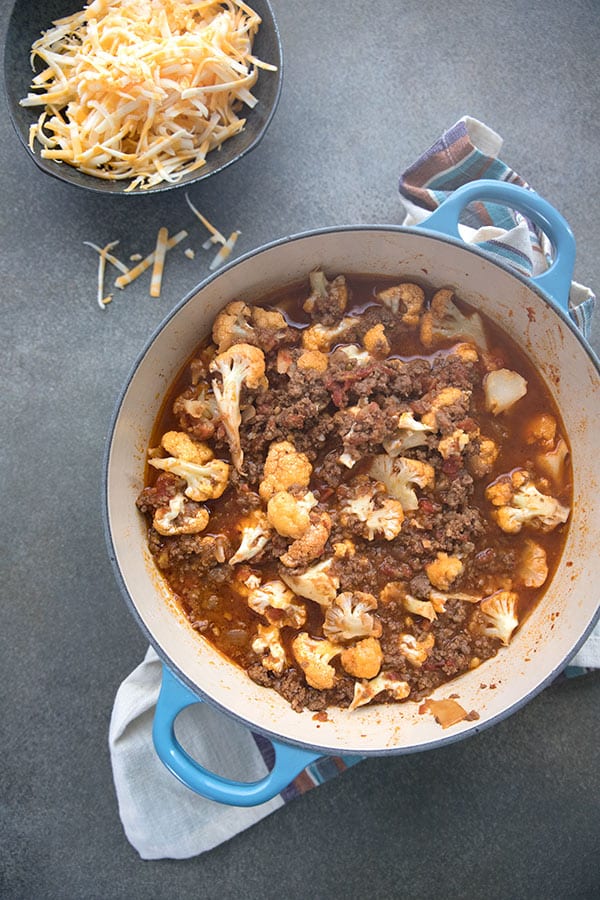 A large blue dutch oven with chili and cauliflower, and a bowl of grated cheese off to the side.