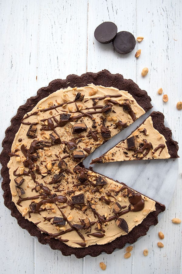 Top down photo of a low carb peanut butter dessert pizza with a slice taken out of it, on a white table. Sugar-free peanut butter cups and peanuts are scattered around. 