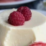 Titled image of White Chocolate Keto Panna Cotta on a white plate with two fresh raspberries on top.