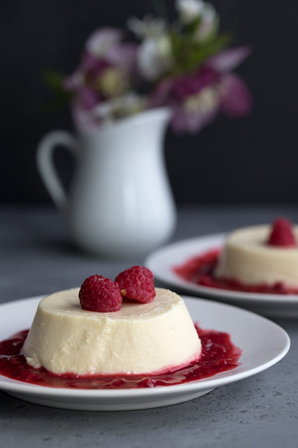 Two plates of panna cotta with raspberry sauce on a grey table, with flowers in the background. 