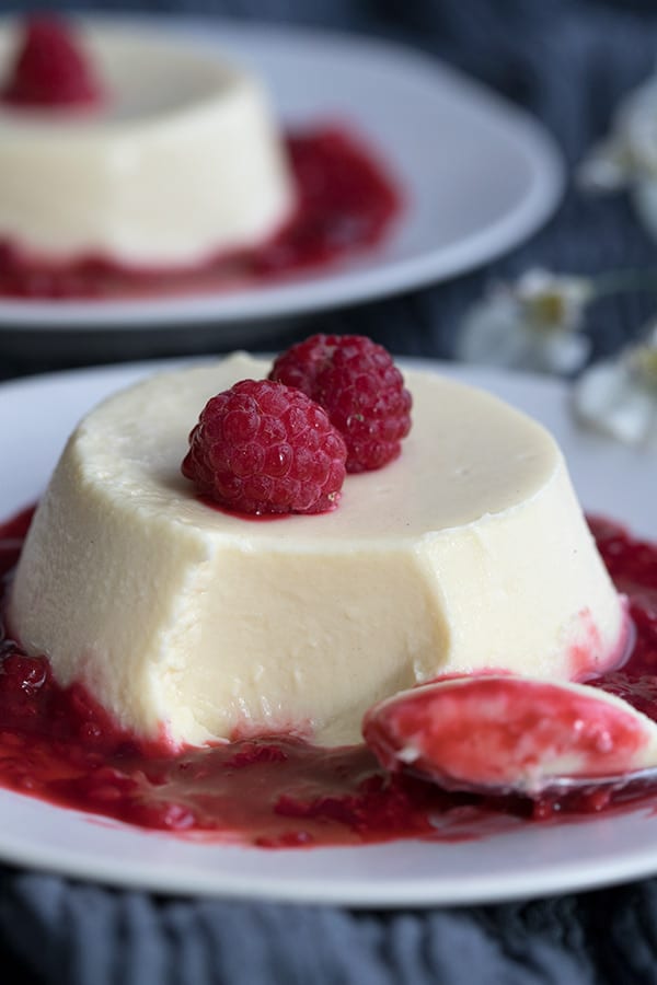 Close up of keto panna cotta with a spoonful taken out of it. Raspberry sauce around the base of the panna cotta and a fresh raspberry on top. 