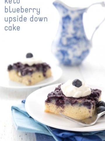 Two slices of blueberry upside down cake on white plates on a white table. A blue patterned jug sits in the background.