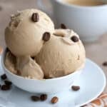 Two scoops of Keto Coffee Ice Cream in a white bowl with coffee beans sprinkled on top and a cup of coffee in the background.