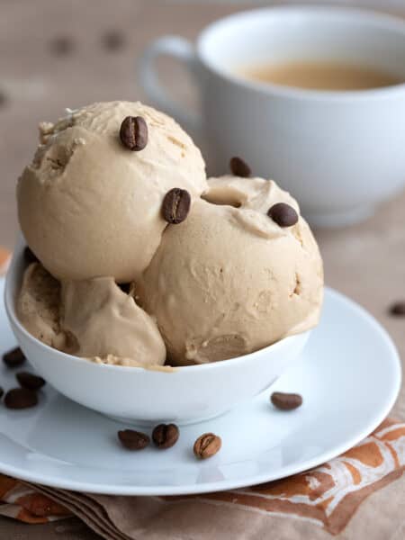 Two scoops of Keto Coffee Ice Cream in a white bowl with coffee beans sprinkled on top and a cup of coffee in the background.