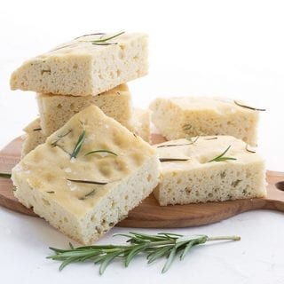 Slices of keto focaccia on a small wooden cutting board. A sprig of rosemary in front.