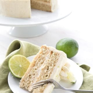 A slice of margarita cake on a white plate over a green napkin, with limes and the rest of the cake in the background.