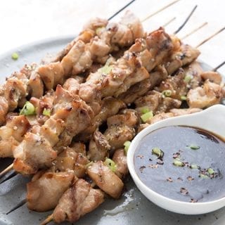 Titled image of keto chicken yakitori skewers on a grey plate with a white dish of yakitori sauce