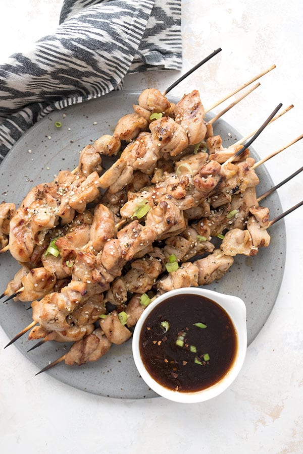 Top down photo of keto chicken skewers on a grey plate with a grey patterned napkin and a dish of yakitori sauce.