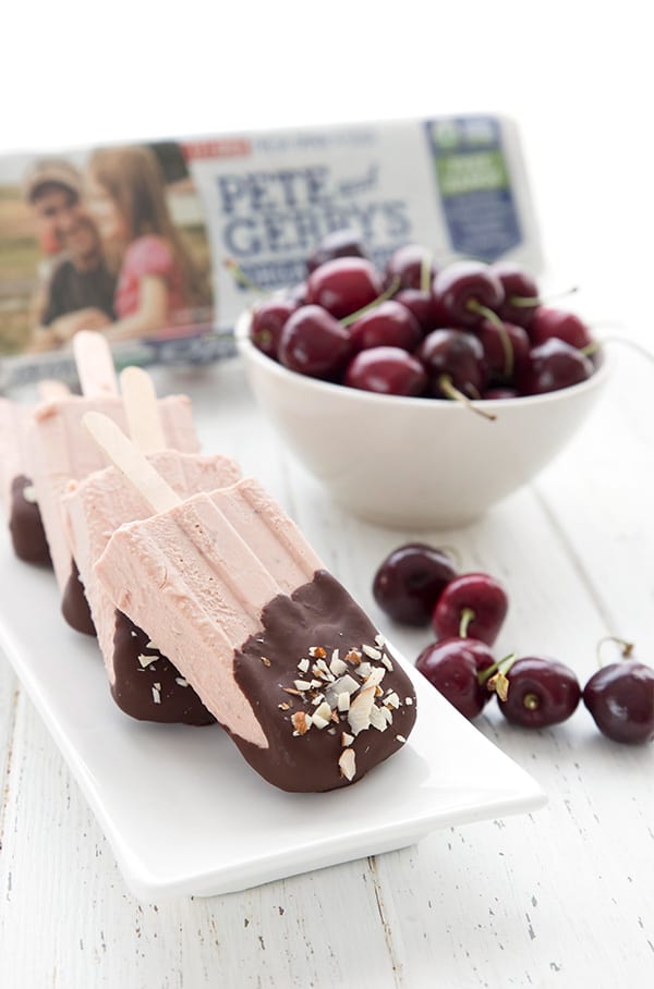 Low carb cherry popsicles dipped in dark chocolate, on a white platter. A bowl of cherries and a carton of eggs are in the background.
